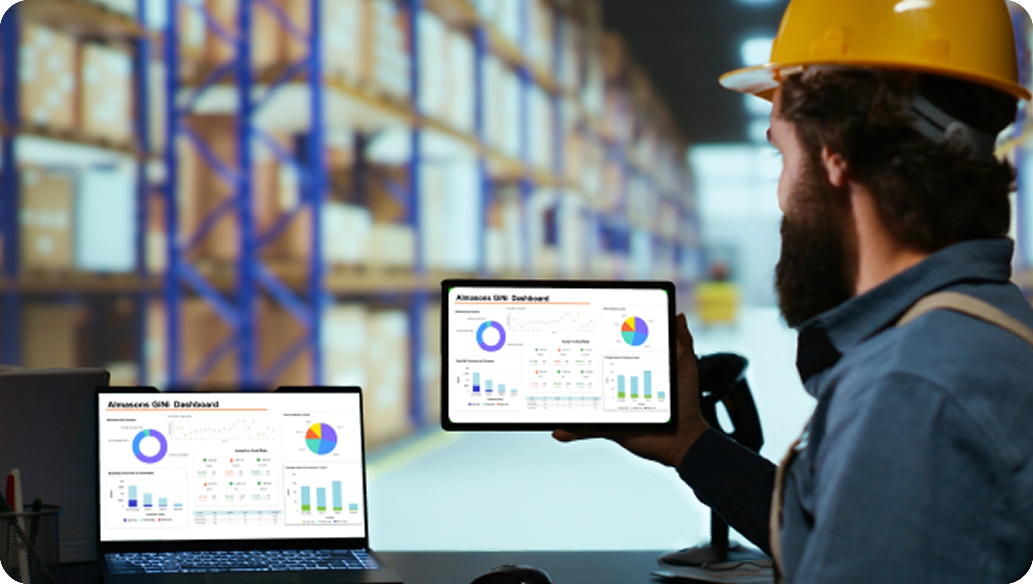 A warehouse worker in a yellow hard hat reviews analytics dashboards on a laptop and tablet, with shelves stocked with boxes in the background. The dashboards display charts and graphs, conveying efficiency and data‑driven operations. 