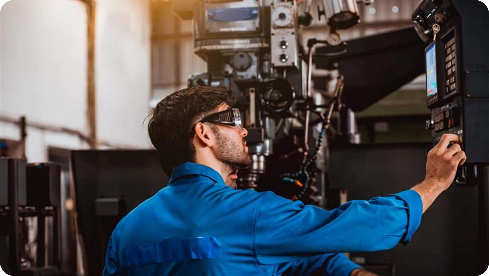 A man in a blue jumpsuit and safety glasses operates a complex machine in an industrial setting, focusing intently on the control panel.