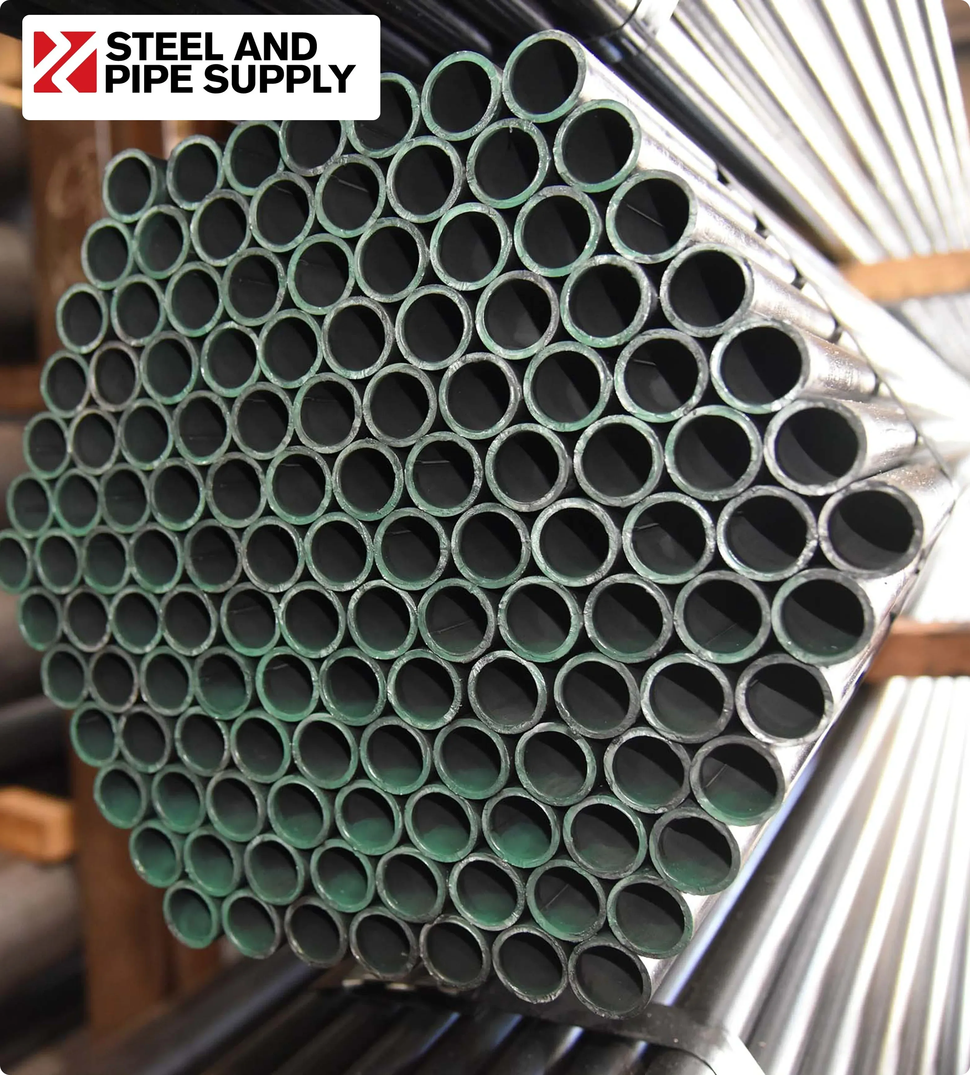 A close-up view of stacked steel pipes with green ends, forming a hexagonal shape. The Steel and Pipe Supply logo is in the upper left corner.